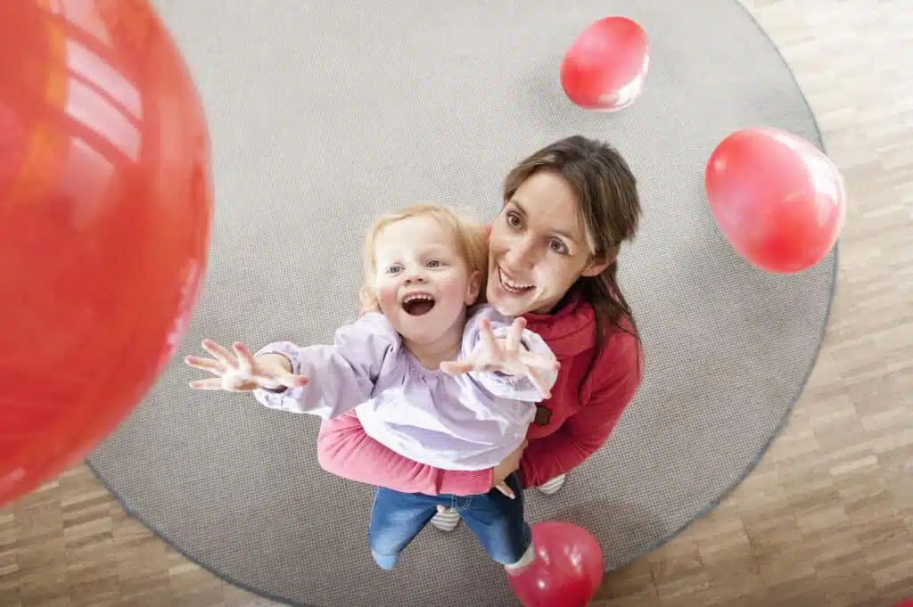 Female educator and little girl playing with red balloons in kindergarten